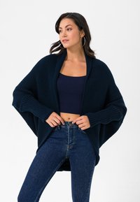 Navy suuruses kudum, millel on laiad varrukad, ribatud tekstuur ja avatud esikülg, kombineeritud tumesinise lühikese topi ja kõrge talje ning teksadega.