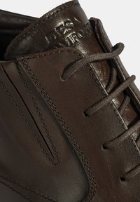 Chaussure en cuir brun avec des lacets foncés, présentant une finition texturée, un logo embossé sur la languette et un design classique à lacets.