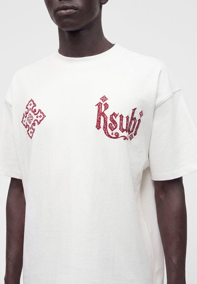 Μαύρο άτομο που φοράει λευκό t-shirt με κόκκινο κεντημένο διακοσμητικό κείμενο "Ksubi" και ένα διακοσμημένο έμβλημα στο πάνω μέρος του στήθους.
