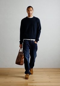 Navy ribbed sweater, white shirt, dark jeans, brown bag, suede shoes. Model stands on wooden floor with light gray background.