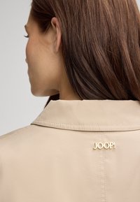 Giacca beige con colletto, caratterizzata da un accento dorato con il logo "JOOP!" sulla parte posteriore. Tessuto liscio e vestibilità su misura.