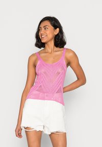 Top in maglia di un brillante colore rosa con un motivo a zigzag, caratterizzato da sottili spalline e un design a trama aperta. Abbinato a pantaloni corti bianchi strappati.