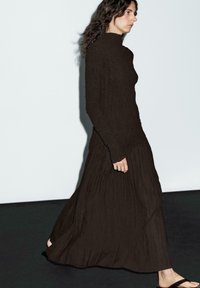 Long, dark brown dress with high neck, textured fabric, and pleated skirt. Paired with flat sandals. Model walking against a light background.