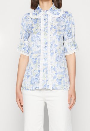 Femme portant un chemisier bleu clair à motif floral avec bordure blanche à volants, manches mi-longues bouffantes et col arrondi, associé à un pantalon blanc.
