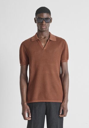 Joven con grandes gafas de sol negras y una camisa tipo polo de punto marrón de manga corta, de pie frente a un fondo liso y claro.