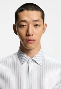 Hvid skjorte med knapper, der har lodrette blå striber, klassisk krave og glat stoftekstur. Modellen har kort hår og en minimal ansigtsudtryk.