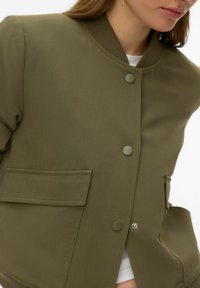 Veste vert olive avec col côtelé, boutons-pression et poches latérales. Présente une texture lisse et une coupe légèrement cintrée.