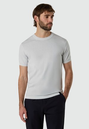 Uomo con barba che indossa una maglia a maniche corte in maglia grigio chiaro e pantaloni scuri, in piedi con una mano in tasca, che guarda verso destra.