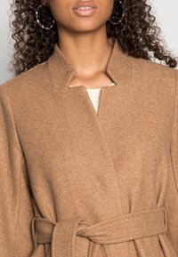 Manteau en mélange de laine couleur fauve avec col châle, taille ceinturée et texture douce, présentant des coutures minimales et un design épuré.