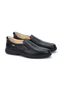 Un par de zapatos slip-on para hombre de cuero negro con plantillas acolchadas y suelas de goma, que exhiben una pequeña etiqueta de marca en el lateral.