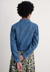 Veste en denim bleu moyen avec un col, des manches longues et une taille cintrée. Comprend des poches et des poignets boutonnés, associée à une jupe à fleurs.