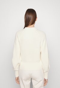 Donna con lunghi capelli castani che indossa un maglione in maglia crema con polsini e orlo a coste, e pantaloni chiari, di fronte a uno sfondo bianco semplice.