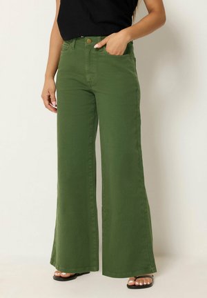 Mujer con pantalones anchos de talle alto color verde oliva con bolsillos delanteros y una camiseta negra sin mangas, de pie frente a un fondo liso.