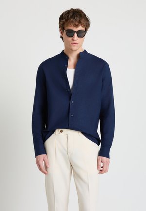 Junger Mann mit schwarzer Sonnenbrille, dunkelblauem Hemd über einem weißen Oberteil und cremefarbenen Hosen, der vor einem einfarbigen Hintergrund steht.