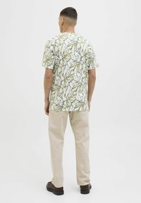 Cămașă cu mânecă scurtă, cu un fundal alb și un model de frunze în verde, albastru și galben. Asortată cu pantaloni bej și pantofi maro închis.