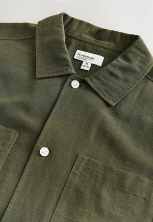 Olijfgroene blouse met een kraag, twee borstzakken en witte knopen. Gemaakt van een gestructureerde stof met horizontale lijnen.