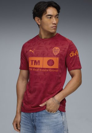 Rød kortærmet trøje med geometrisk mønster, prydet med orange sponsorlogoer og et våbenskjold. Fremstillet af letvægtsstof med en glat tekstur.