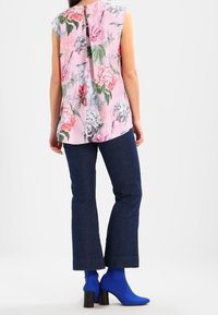 Blusa floral en rosa con mangas cortas y abertura en la parte posterior, combinada con jeans de campana azul oscuro y botas de tobillo azules.