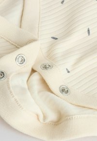 Tissu côtelé couleur crème avec des boutons-pression argentés partiellement attachés, montrant un tricot texturé et de petits détails sombres cousus.