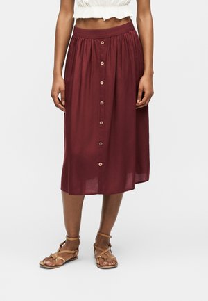 Femme portant une jupe midi bordeaux avec des boutons sur le devant, associée à des sandales plates à brides couleur cuir clair et un top court blanc.