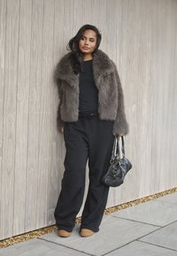 Szara kurtka z sztucznego futra na czarnej bluzce, w zestawie z czarnymi spodniami w szerokim kroju. Trzyma czarną torebkę, stojąc przy drewnianej ścianie.