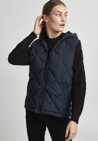 Navy blue quilted vest with a hood, featuring a front zipper, side pockets, and textured black knit sleeves with a cable pattern.