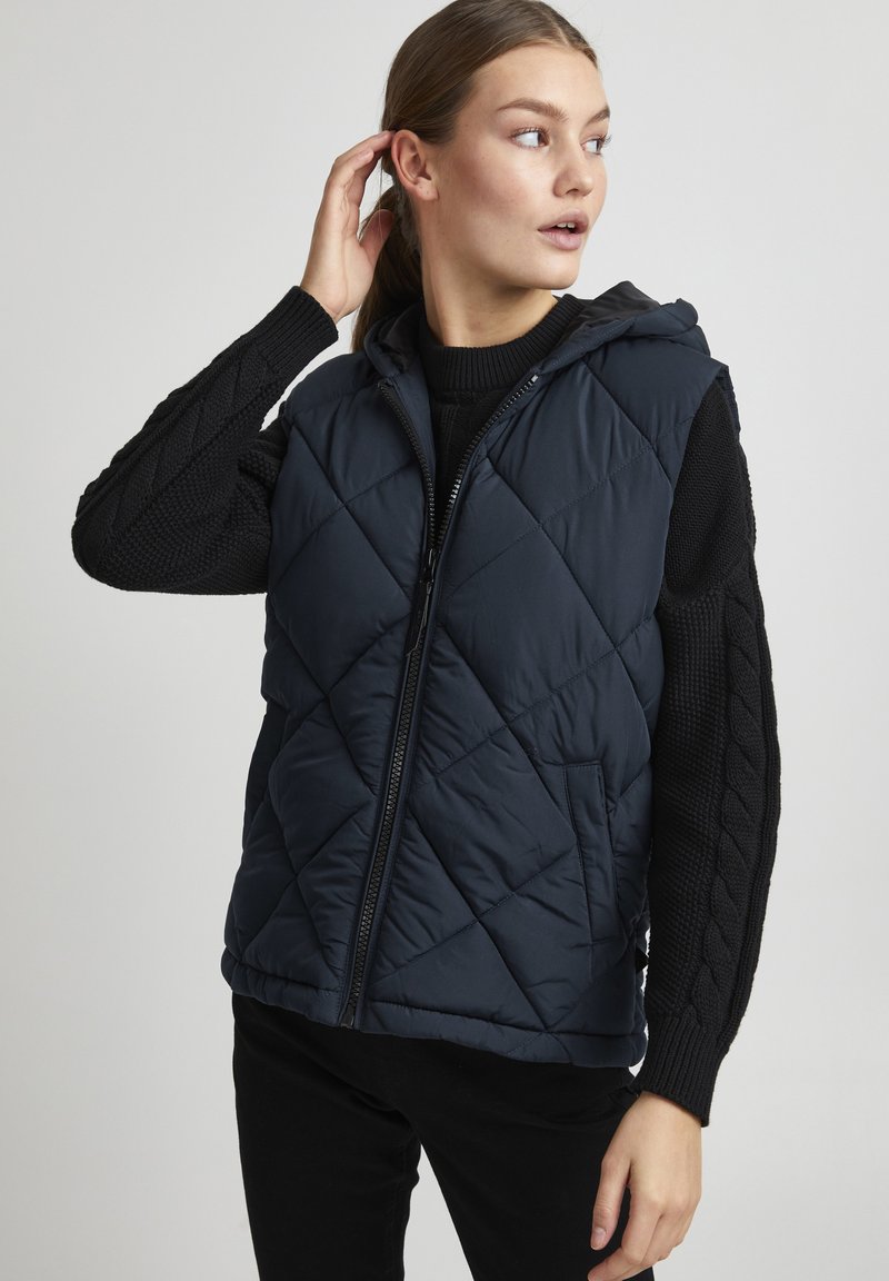 Navy blue quilted vest with a hood, featuring a front zipper, side pockets, and textured black knit sleeves with a cable pattern.