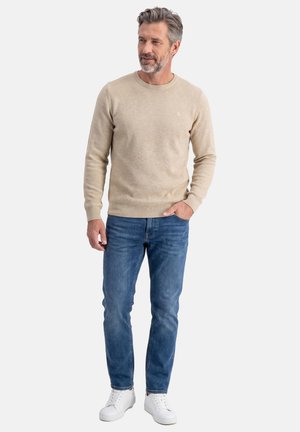 Homme d'âge moyen debout, portant un pull beige, un jean bleu et des baskets blanches, avec une main dans la poche, sur un fond uni.