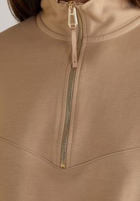 Jersey color canela con cuello alto, que presenta una cremallera dorada y tela texturizada. El diseño incluye detalles de costuras a lo largo del frente.