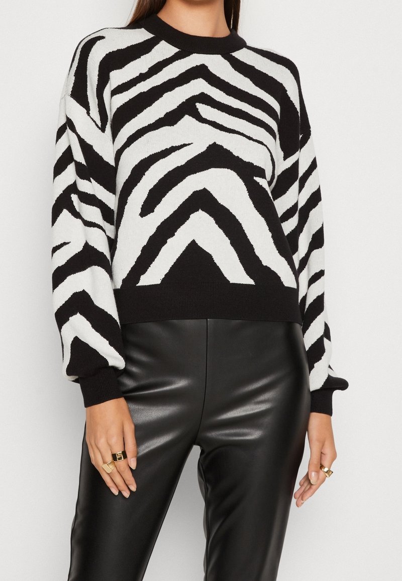 Femme portant un pull à motif zèbre noir et blanc et un pantalon en cuir noir, avec des bagues dorées aux deux mains.