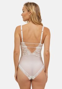 Body en dentelle blanche avec un dos en mesh transparent, des détails en dentelle élaborés, des bretelles ajustables et un nœud décoratif en bas.