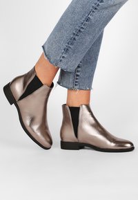 Metallinhohtoiset pronssinväriset nilkkurit joustavilla sivuilla, pyöreällä kärjellä ja mustalla litteällä pohjalla; sileä pinta ja tyylikäs muotoilu.