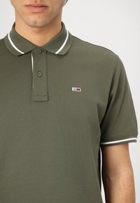 Olivovo zelené polo tričko vyrobené z textúrovanej látky. Má golier s bielymi pruhmi a malým farebným logom na hrudi.