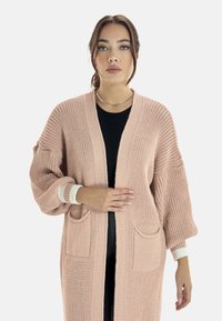 Lång, mjuk stickad cardigan i ljusrosa, med överdimensionerade ärmar, framfickor och kontrasterande ribbad kant i vitt.
