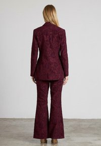 Bordeaux gekleed met een bloemenpatroon met een op maat gemaakte blazer en uitlopende broeken van textuurstof. Zicht van achteren waarbij het ontwerp wordt benadrukt.