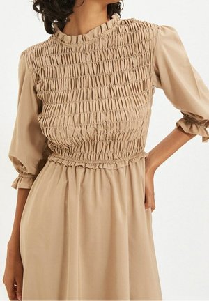 Beige jurk met een gerimpeld lijfje, driekwart pofmouwen met geplooide manchetten en een gerimpelde hoge halslijn, gedragen met één hand in de zij.