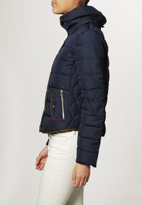 Marineblaue, gesteppten Pufferjacke mit tailliertem Schnitt, hohem Kragen, braunen Akzenten und einer Reißverschlusstasche an der Seite.