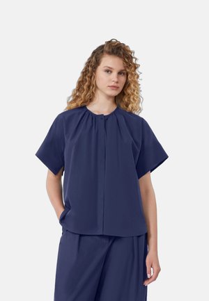 Donna con capelli ricci che indossa una maglietta blu scuro a maniche corte ampia e pantaloni larghi abbinati, in piedi con una mano in tasca.