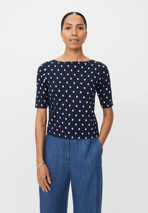 Granatowa bluzka w groszki z krótkimi rękawami, wykonana z miękkiego materiału. Posiada zaokrąglony dekolt i prosty krój, zestawiona z jasnymi dżinsowymi spodniami.