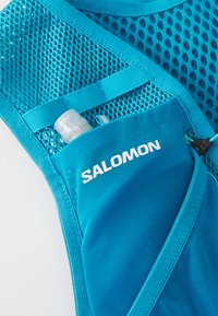 Kék Salomon nejlon futó mellény, amelynek elülső zsebében egy kis átlátszó vízpalack található, fehér háttér előtt.