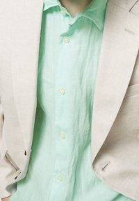 Jasny lniany żakiet w połączeniu z miętowo-zieloną koszulą z guzikami. Koszula ma miękki, przezroczysty materiał i okrągłe guziki.