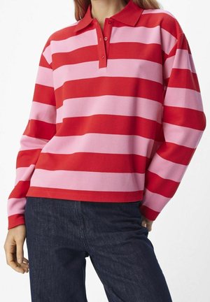 Mujer vistiendo una camiseta polo de manga larga con anchas rayas horizontales rojas y rosas, combinada con vaqueros azul oscuro.