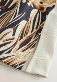 Blomsterpatron stof med beige og brune nuancer, med en struktureret overflade. Kantstikking synlig på en lysfarvet søm.