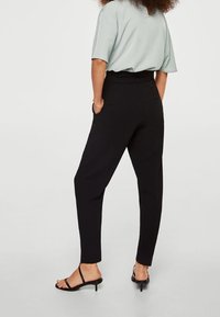 Pantaloni neri a tapered con una texture liscia e una vita aderente, abbinati a una maglietta verde chiaro a maniche corte e sandali neri.