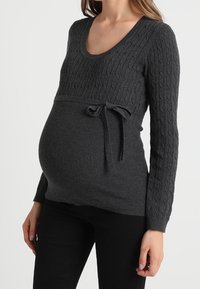 Ciemnoszary dzianinowy sweter ciążowy z prążkowaną teksturą, wzorem warkocza na górze oraz ozdobnym wiązaniem z boku nad brzuchem.