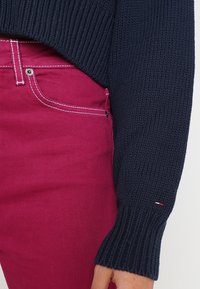 Marineblauer Strickpullover mit gerippten Bündchen, kombiniert mit pinkfarbenen Jeans aus Denim, die kontrastierende weiße Nähte und ein kleines Logo-Detail aufweisen.