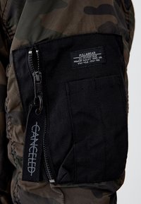 Veste camouflage avec une poche en tissu noir équipée d'une fermeture éclair. L'étiquette de la poche affiche la marque et les instructions d'entretien ; "CANCELLED" est brodé sur la poche.