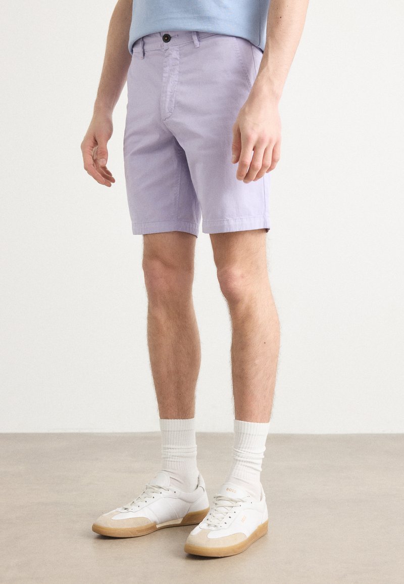 Pantalones cortos morados claros de algodón, con cierre de botón y dos bolsillos laterales. Combinados con zapatillas blancas y calcetines blancos de caña baja.