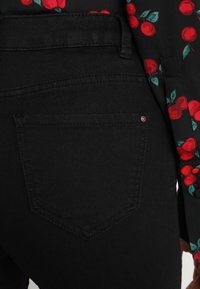 Pantalones de denim negro con un bolsillo trasero que presenta un sutil detalle de costura. La tela tiene una textura suave, combinada con una camisa con un estampado floral.