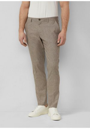 Mężczyzna w wąskich, jasnobrązowych teksturowanych spodniach i białych trampkach, stoi na tle jednolitego, jasnoszarego tła.